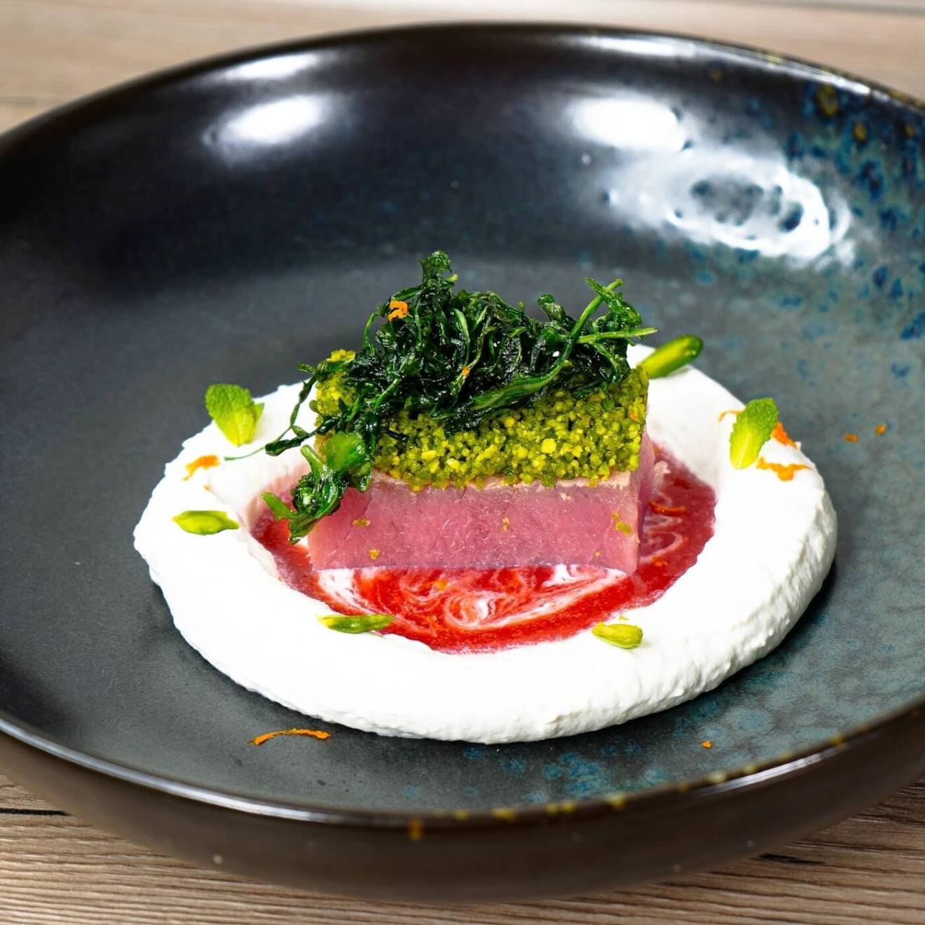 Colorful gourmet dish with fish, green topping, sauce, and cream served on a dark plate.