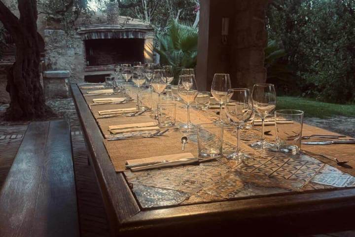 Outdoor dining table with glasses under warm lighting, surrounded by trees and brick path.
