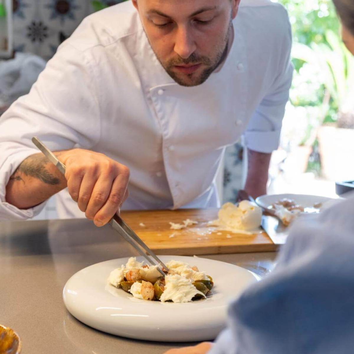 Chef plating a dish with tweezers while a person watches on in a kitchen.