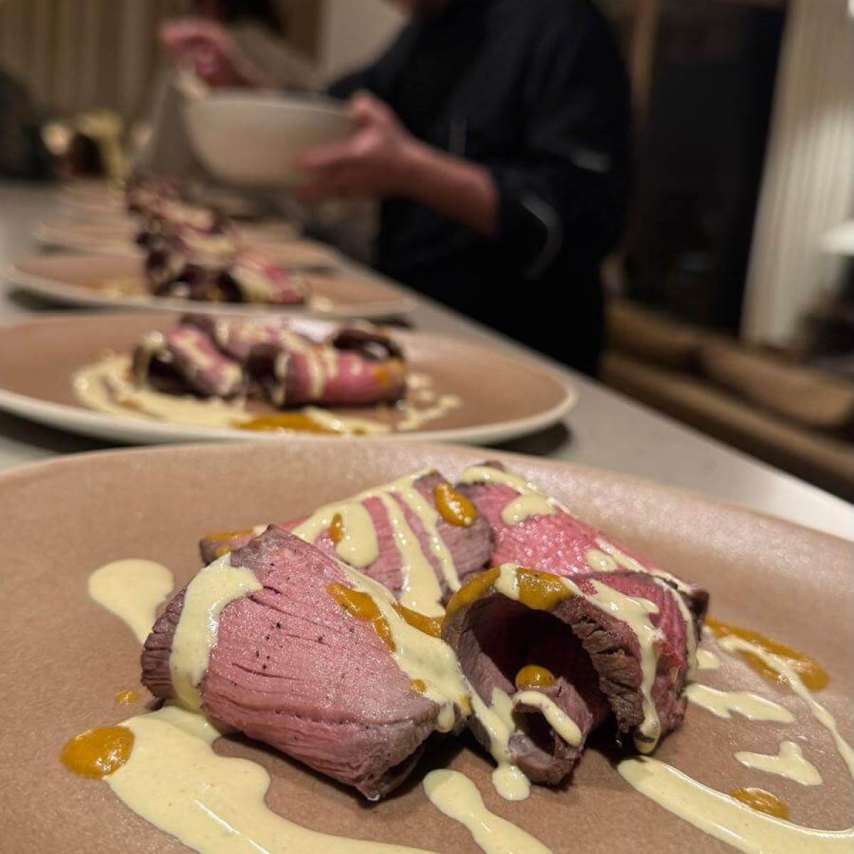 Sliced meat with creamy sauce on plate; chef preparing dish in background.
