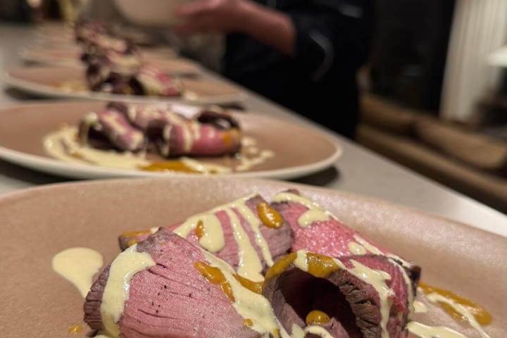 Sliced meat with creamy sauce on plate; chef preparing dish in background.