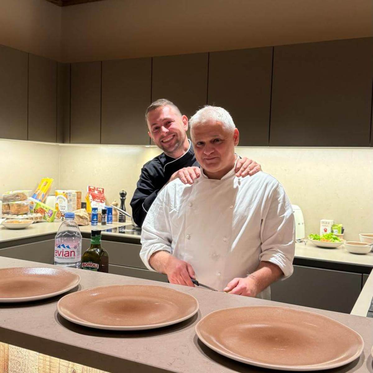 Two men in a kitchen: one in chef attire, the other in a black shirt, with four empty plates on the counter.