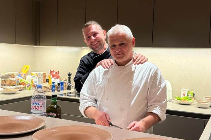 Two men in a kitchen: one in chef attire, the other in a black shirt, with four empty plates on the counter.