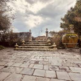 Stone steps lead to a garden path with large pottery and bushes under a cloudy sky.