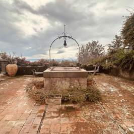 Terracotta tiled terrace with an old well, surrounded by lush greenery and cloudy sky.