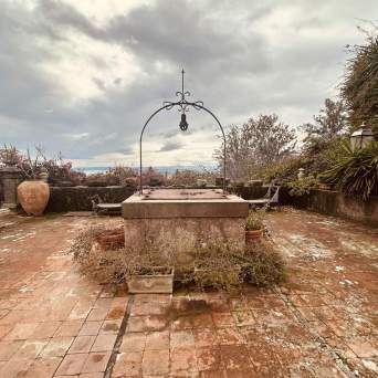 Terracotta tiled terrace with an old well, surrounded by lush greenery and cloudy sky.