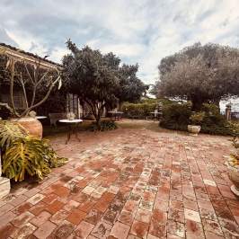 Brick patio with trees, plants, tables, and stone urns under a cloudy sky.