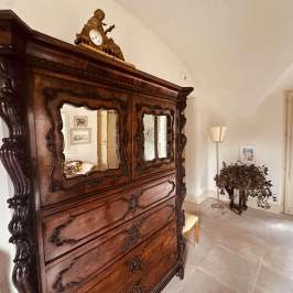 Room with antique wooden dresser, ornate mirrors, clock on top, plant, and lamp nearby.