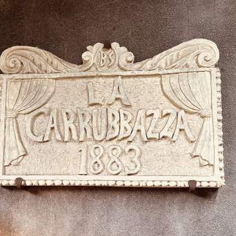 Ornate stone sign with 'La Carrubazza 1883' carved, featuring curtain design.