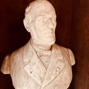 Stone bust of a man with a bow tie and beard against a dark background.