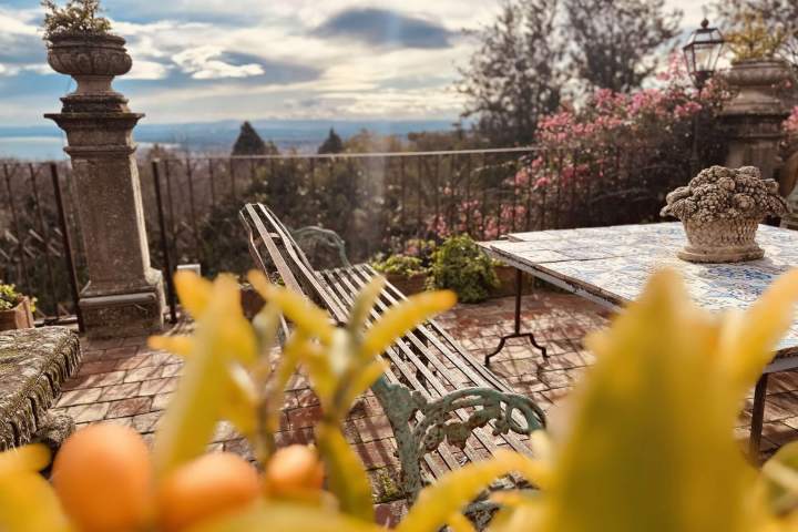 Sunny patio with metal bench, tiled table, and plants, overlooking landscape with trees and distant view.