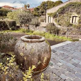 Large stone pot in garden with cobblestone path, greenery, and rustic house in background.