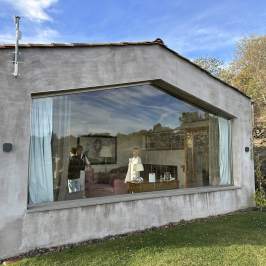 Modern house with large window reflecting scenery and two people inside, surrounded by grass and trees.