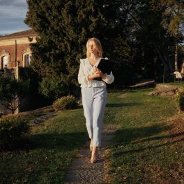 Woman in white outfit walking on garden path, holding a book, near a house.