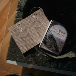 Two wine glasses on a mat beside a book titled 'ETNA WINE' on a stone table.