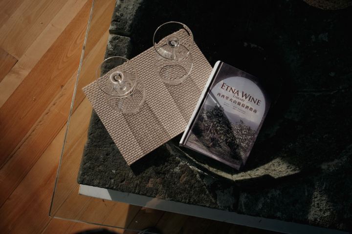 Two wine glasses on a mat beside a book titled 'ETNA WINE' on a stone table.