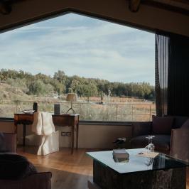 Room with large window showing forest view, desk, chair, armchairs, and a glass table.