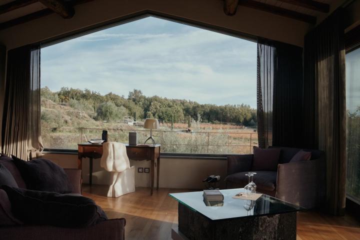 Room with large window showing forest view, desk, chair, armchairs, and a glass table.