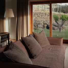 Cozy room with brown sofa, floor lamp, large window showing garden, stone wall and tree.