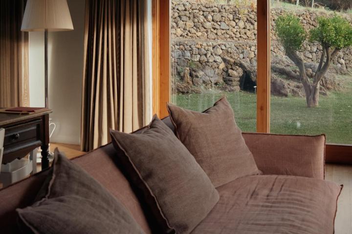 Cozy room with brown sofa, floor lamp, large window showing garden, stone wall and tree.