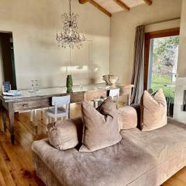 Cozy living room with tan sofa, wooden table, chandelier, and large window with view of garden.