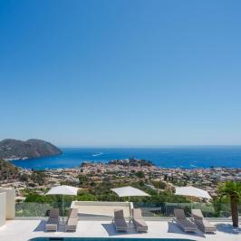 Poolside view with loungers, umbrellas, hillside town, and ocean in background under clear blue sky.