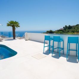 Poolside with blue chairs, palm tree, and sea view, under clear blue sky.