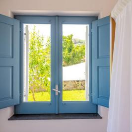 Open window with blue shutters overlooks green garden and tree outside.