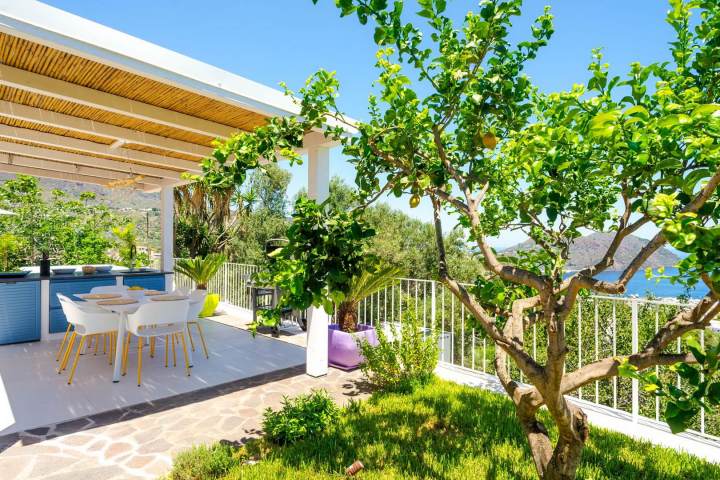 Patio with white chairs, a dining table, trees, and sea view under a pergola on a sunny day.