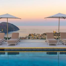 Poolside loungers and umbrellas overlooking a coastal town and sea at sunset.