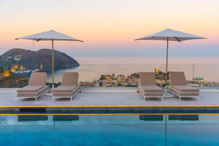 Poolside loungers and umbrellas overlooking a coastal town and sea at sunset.