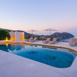 Infinity pool with sun loungers and umbrellas overlooking sea and hills at sunset.