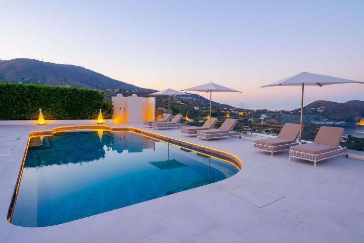 Infinity pool with loungers and umbrellas at sunset, overlooking mountains and sea.