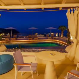 Poolside seating with curtains, round table, and chairs overlooking a pool at night with umbrellas and palm tree.