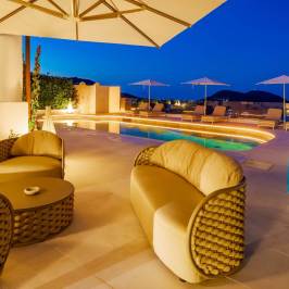 Outdoor lounge area with sofa, chairs, tables, pool, and umbrellas at night with ambient lighting.