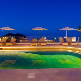 Rooftop pool at night with umbrellas, loungers, and a palm tree under a deep blue sky.