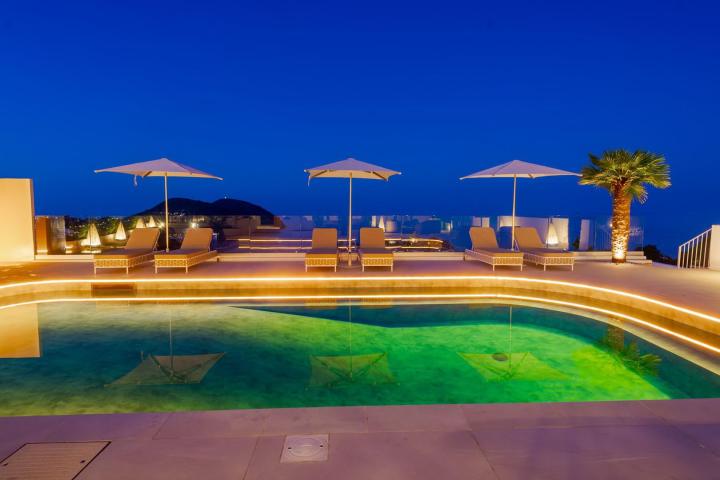 Rooftop pool at night with umbrellas, loungers, and a palm tree under a deep blue sky.