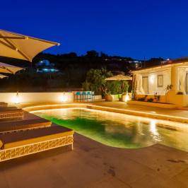 Outdoor pool area at night with loungers, umbrellas, and a lit villa.
