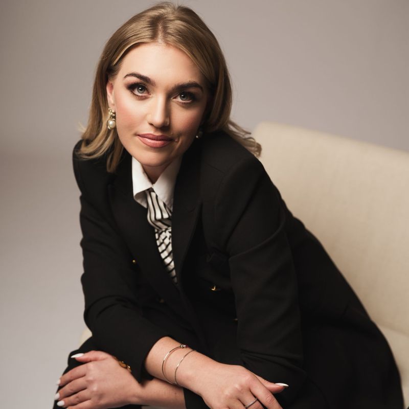 Woman in black suit sitting on chair, looking confidently forward.