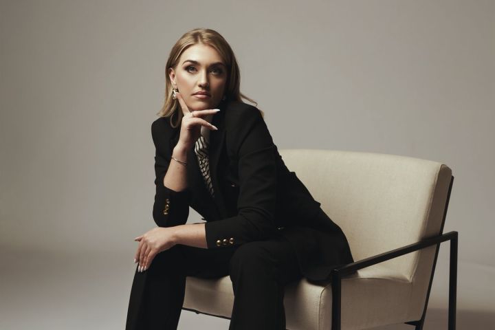 Woman in a black suit sitting on a beige chair in a minimalist setting.