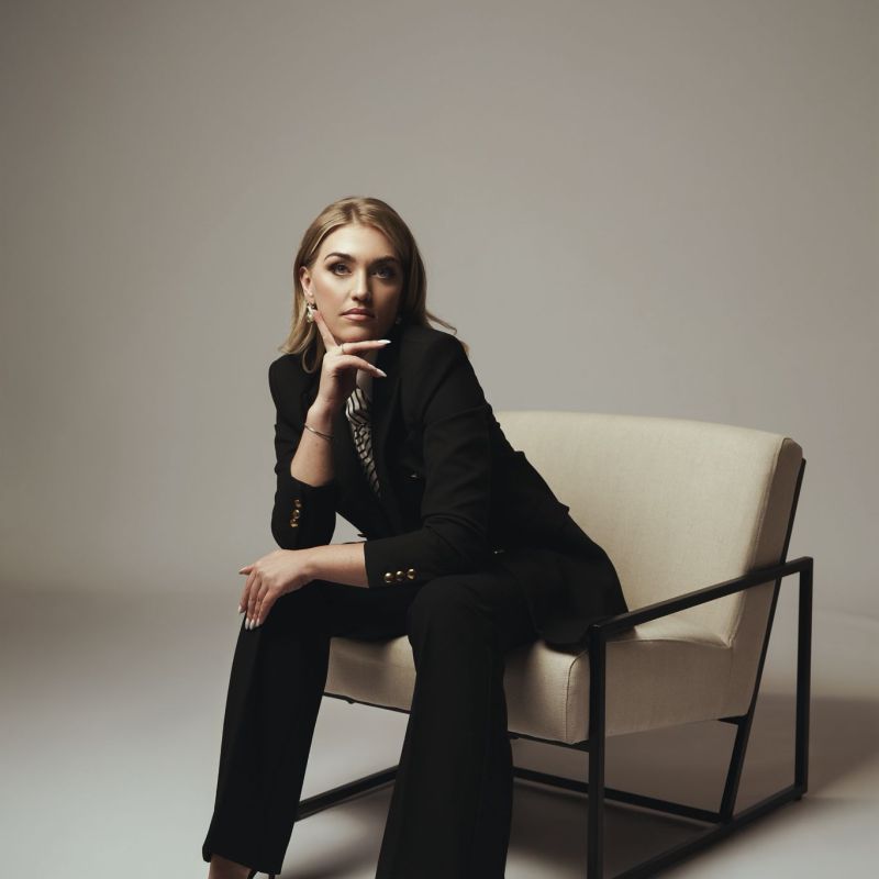Woman in a black suit sitting on a beige chair in a minimalist setting.