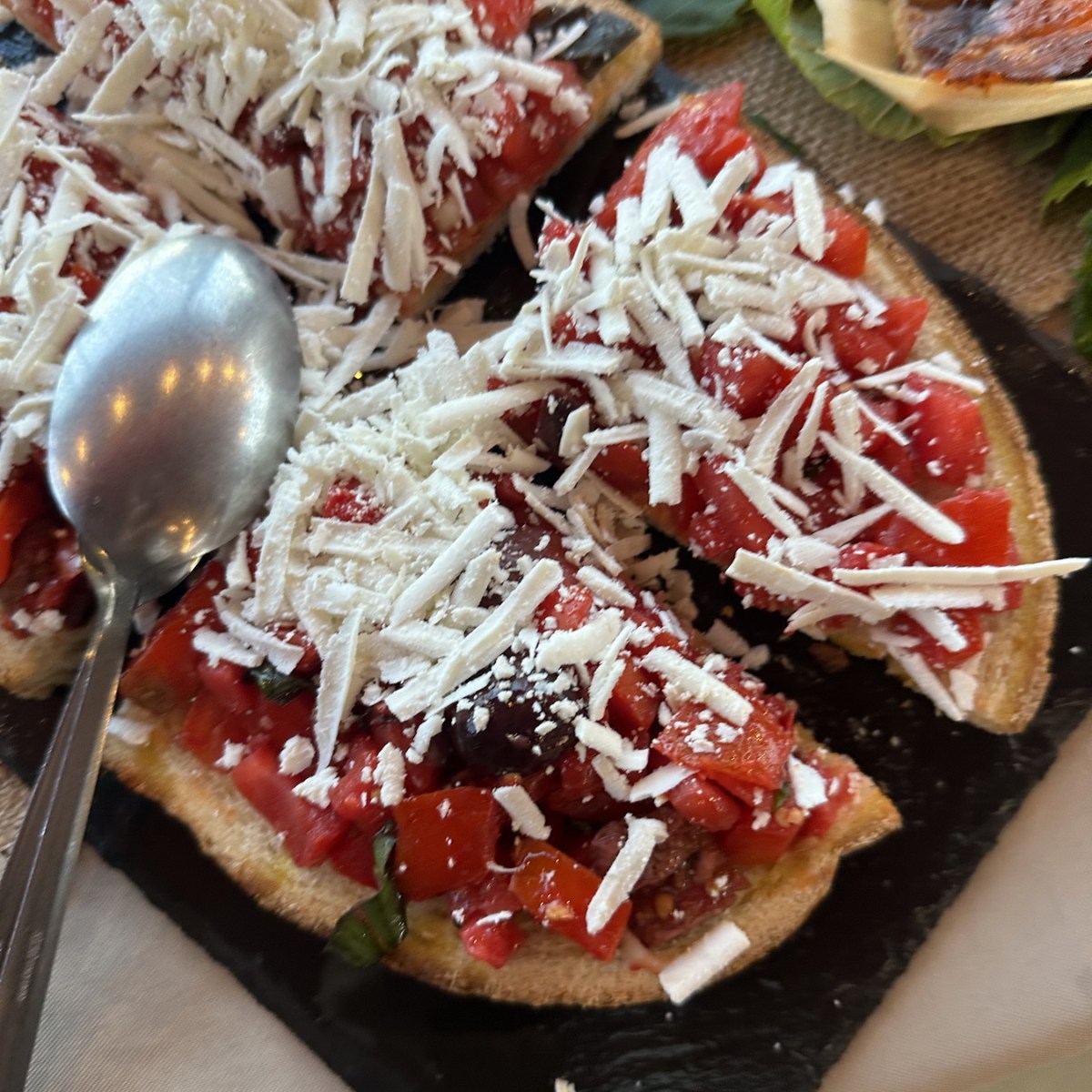 Sliced bruschetta topped with chopped tomatoes and grated cheese on slate, with a spoon on the side.