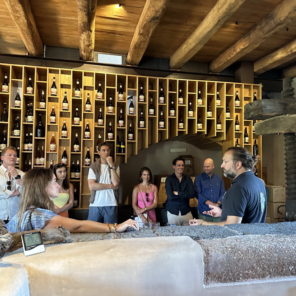 Group listening to a speaker in a wine cellar with wooden beams and bottles displayed on shelves.