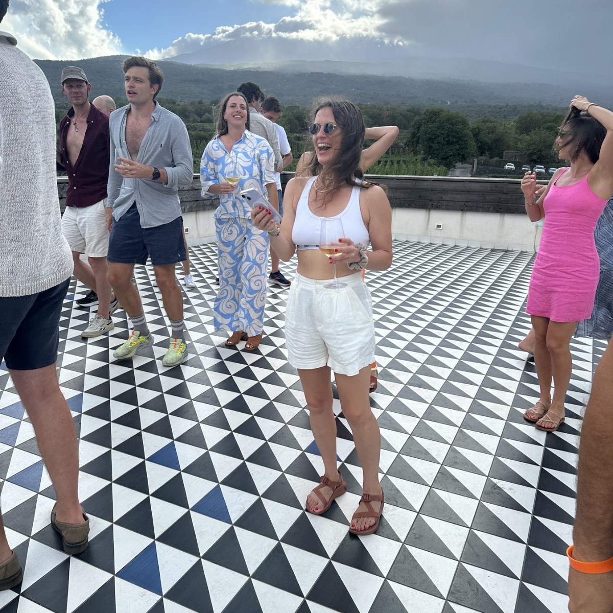 People on patterned terrace with drinks, cloudy sky in background.