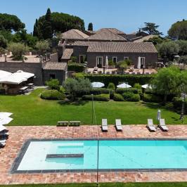 Aerial view of a pool with loungers in front of a villa surrounded by trees and shrubs.