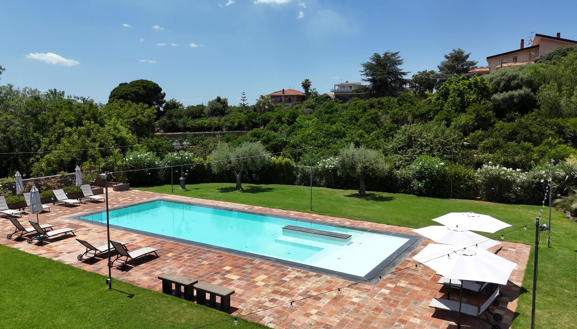 Rectangular pool with loungers and umbrellas on a sunny day, surrounded by green landscape.