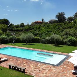 Rectangular pool with loungers and umbrellas on a sunny day, surrounded by green landscape.