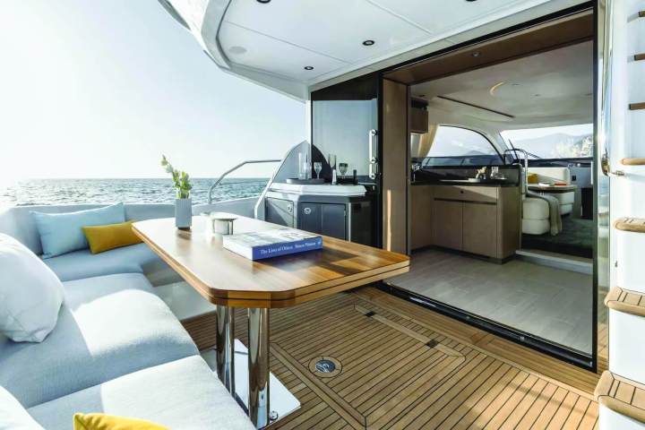 Elegant yacht with outdoor seating, wooden table, and open sliding doors to cabin, overlooking the sea.